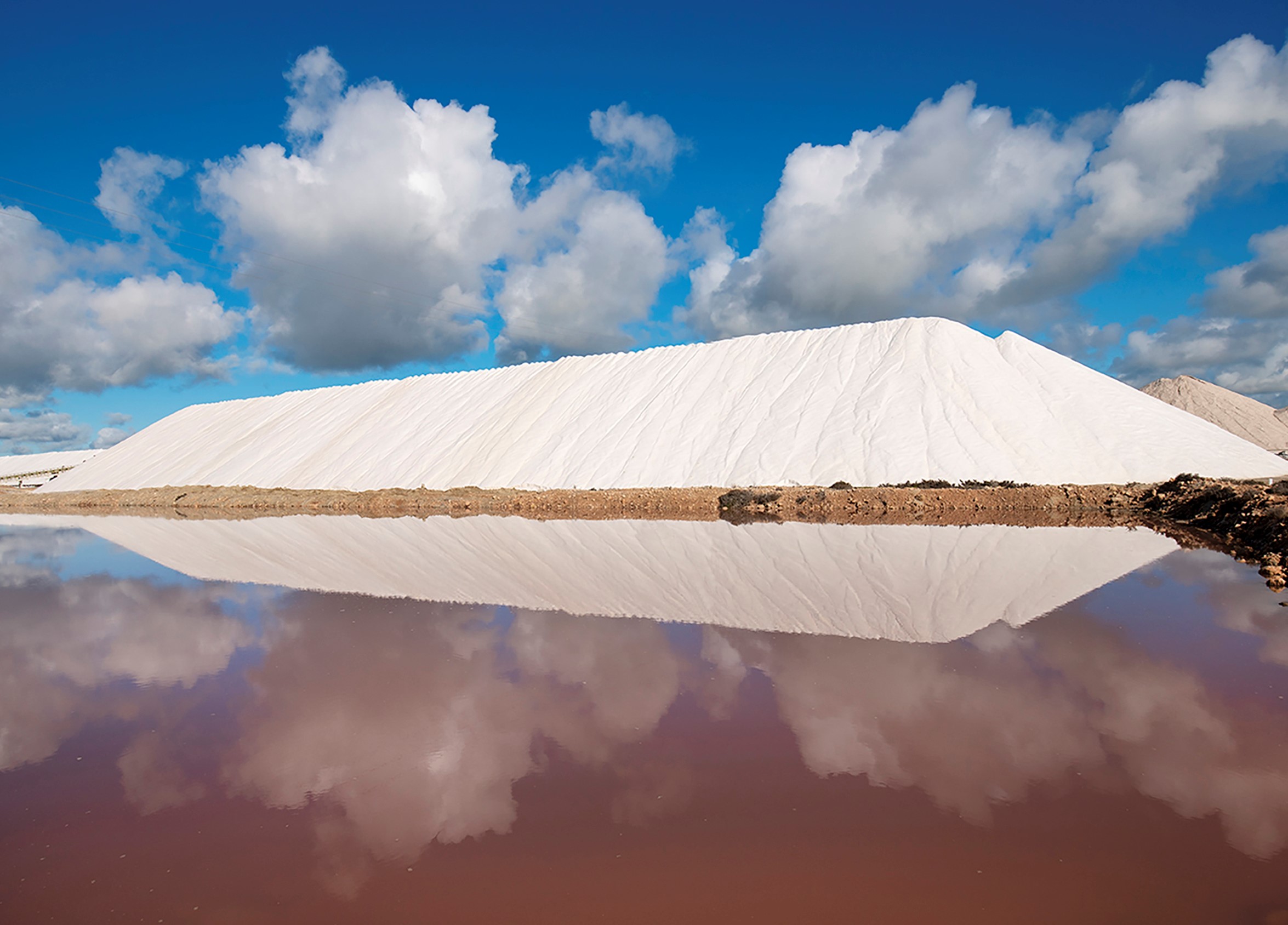 A photo of a salt pile with a bright blue sky and the clouds reflecting in pink sea water